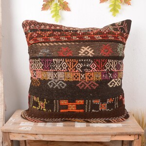 Puede incluir: Cojín decorativo cuadrado con un diseño tejido. El cojín presenta una mezcla de tonos marrones, rojos, naranjas y morados. Se coloca sobre una superficie de madera, mostrando sus intrincados detalles y texturas.