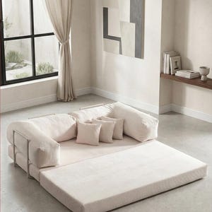 May include: A modern, modular sofa in a neutral color palette. The sofa features a low profile with a large, flat base and cushioned backrests and armrests. Small square pillows add comfort. The design includes metal supports and a light-colored rug.