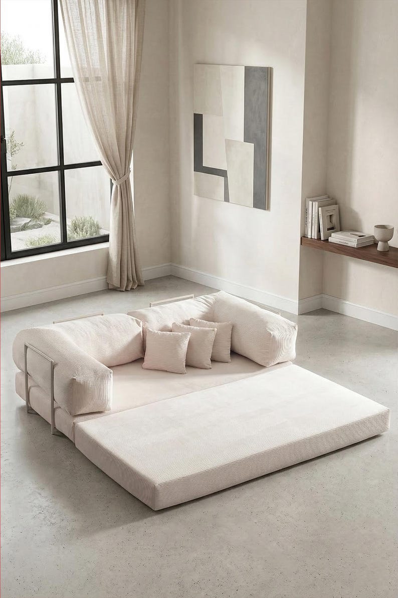 May include: A modern, modular sofa in a neutral color palette. The sofa features a low profile with a large, flat base and cushioned backrests and armrests. Small square pillows add comfort. The design includes metal supports and a light-colored rug.