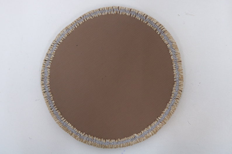 May include: A round, brown fabric patch with a light gray and beige stitched border.