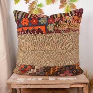 May include: A decorative square pillow with a patchwork design. The top and bottom sections feature colorful, patterned fabric, while the center has a geometric design. The pillow is placed on a wooden surface, suggesting a home decor item.