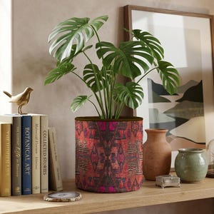 May include: A cylindrical plant pot featuring a vibrant red and dark patterned textile exterior with geometric motifs. The pot contains a green monstera plant with large, split leaves. The design displays intricate, symmetrical dark shapes.