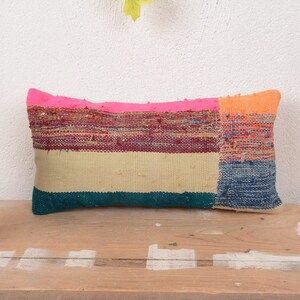 May include: Rectangular decorative pillow with a vibrant, textured design. The pillow features a pink top, an orange right side, and a striped pattern of teal, beige, and burgundy. It rests on a wooden surface.