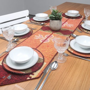 Puede incluir: Una mesa de madera puesta con un mantel individual rojo, naranja y marrón. El mantel individual tiene un patrón geométrico. Hay platos, cuencos y vasos blancos sobre la mesa.