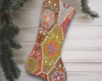 May include: A patterned Christmas stocking in shades of brown, green, orange, and purple. The geometric design is hanging from a silver loop. The background is a white wooden surface, with a pine branch and festive decorations visible.