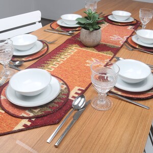 Puede incluir: Una mesa de madera puesta con un camino de mesa estampado en rojo, naranja y verde. La mesa está puesta con cuencos, platos y cubiertos blancos. También hay dos vasos y una pequeña planta en maceta en el centro de la mesa.