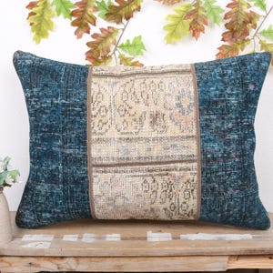 May include: Rectangular cushion with a central panel of beige patterned fabric, framed by teal-coloured fabric. The cushion is placed on a wooden surface, with a small vase of flowers and decorative leaves in the background.