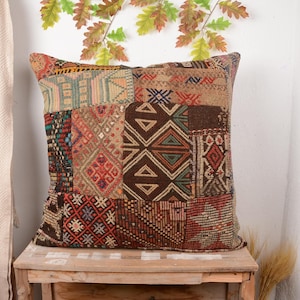 May include: Square patchwork pillow with a variety of geometric patterns in shades of brown, red, and beige. The pillow is placed on a wooden table, with a neutral-colored curtain and decorative leaves in the background.