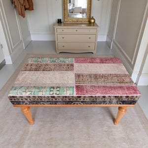 May include: A rectangular ottoman featuring a patchwork upholstery design with sections of teal, cream, brown, and deep red patterned fabric. The piece is supported by four light-toned wooden legs with a turned, tapered silhouette.