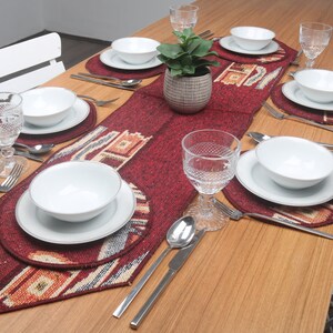 May include: A dining table set for a meal, featuring white bowls and plates, silverware, and patterned placemats and a runner in shades of red, beige, and brown. Clear water glasses and a small potted plant complete the setting.