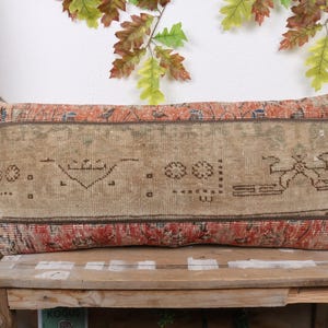 May include: Rectangular beige pillow with a distressed, patterned design. The pillow features a central panel with geometric shapes and borders in red and brown. The pillow is placed on a wooden surface.