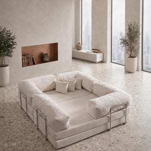 May include: A large, off-white modular sofa with a low profile and a rectangular shape. The sofa features plush, cushioned sections and square pillows. The frame is made of metal. The room has large windows and potted plants, creating a minimalist aesthetic.