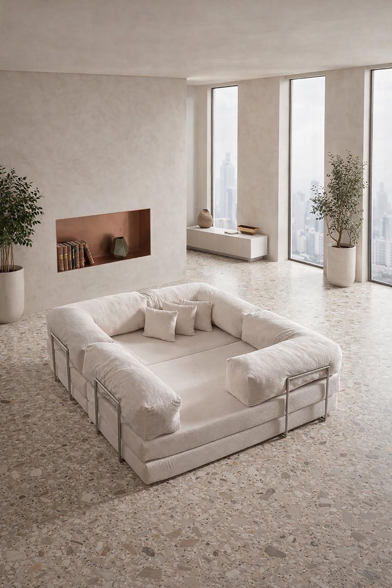 May include: A large, off-white modular sofa with a low profile and a rectangular shape. The sofa features plush, cushioned sections and square pillows. The frame is made of metal. The room has large windows and potted plants, creating a minimalist aesthetic.