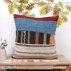 May include: A square decorative pillow with a patchwork design. The top section is blue, followed by a brown and striped section, and a beige and striped bottom. The pillow is resting on a wooden table.