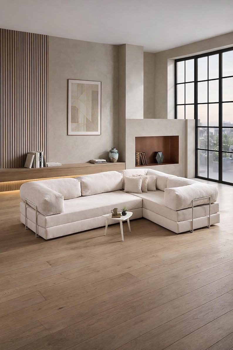 May include: A modern, off-white sectional sofa with a minimalist design. The sofa features a low profile, square arms, and metal accents. It is placed in a neutral-toned living room with a wooden floor and large windows.
