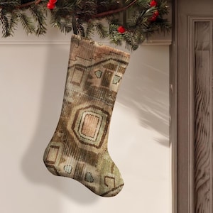 Puede incluir: Un calcetín navideño con un patrón descolorido inspirado en una alfombra en tonos marrón, bronceado, verde apagado y azul suave. El calcetín tiene una forma clásica con un lazo para colgar en la parte superior, mostrando un diseño geométrico.