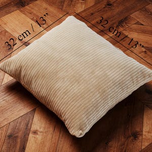 May include: A beige corduroy pillow, measuring 13 inches by 13 inches, and 6 inches thick, rests on a wooden floor. The pillow has a ribbed texture and a square shape. The dimensions are labeled on the image.