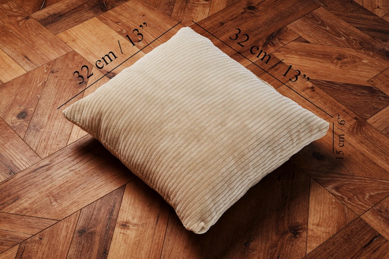 May include: A beige corduroy pillow, measuring 13 inches by 13 inches, and 6 inches thick, rests on a wooden floor. The pillow has a ribbed texture and a square shape. The dimensions are labeled on the image.