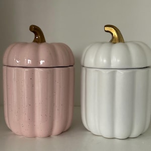 Puede incluir: Dos recipientes de cerámica con forma de calabaza con tapas doradas. Un recipiente es rosa con motas blancas y el otro es blanco.