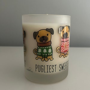 May include: A frosted glass candle with creamy white wax. The candle features cartoon pugs wearing festive sweaters. The text "Pugliest Sweater" is printed on the front.