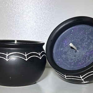 May include: Two black cauldron-shaped candles with white spiderweb designs. One candle is upright, the other tilted, revealing a dark blue wax with glitter. The candles have white wicks, perfect for Halloween or gothic decor.