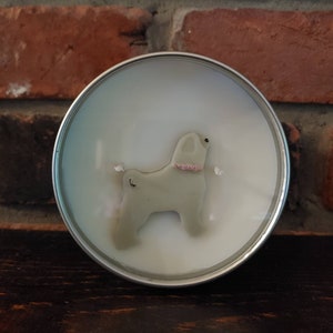 Puede incluir: Vela redonda en lata plateada. La cera presenta un diseño de perro gris claro con collar rosa. Se ven dos mechas. El fondo es una pared de ladrillos rojos. Vela aromática.