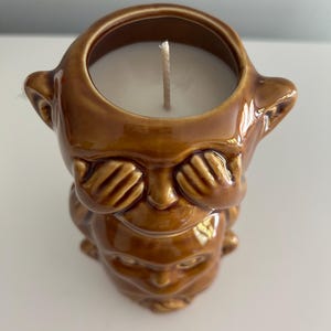 Può includere: Un portacandele in ceramica marrone a forma di tre scimmie, con la scimmia in cima che si copre gli occhi. La candela è bianca e ha uno stoppino.