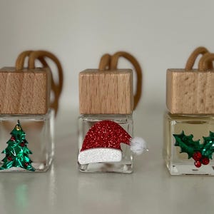 Könnte beinhalten: Drei kleine gläserne Auto-Lufterfrischer mit Holzdeckeln und Schnur-Aufhängern. Die Lufterfrischer sind mit Weihnachtsmotiven verziert: ein grüner Weihnachtsbaum, eine rote Weihnachtsmütze und ein grüner Stechpalmenzweig.