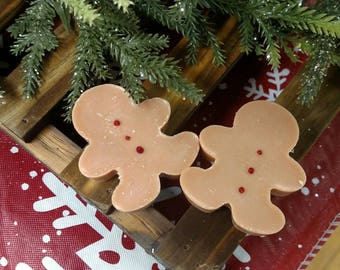 Puede incluir: Dos jabones con forma de hombre de jengibre con chispas de caramelo rojo. Los jabones están sobre una superficie de madera con un patrón de copo de nieve rojo y blanco.