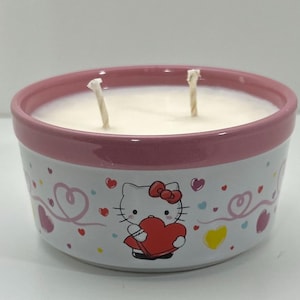 Puede incluir: Una vela redonda en un recipiente de cerámica blanco y rosa. El recipiente presenta un diseño de Hello Kitty con un corazón rojo y corazones decorativos. La vela tiene dos mechas y un relleno de cera blanca.
