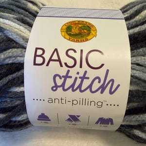 May include: A skein of Lion Brand Basic Stitch anti-pilling yarn featuring a variegated color palette of dark charcoal, medium gray, light gray, and white. The white paper label displays the Lion Brand Yarns logo, the text Basic Stitch, and icons.