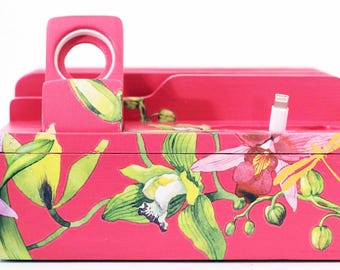 Może przedstawiać: Żyworóżowa drewniana stacja ładująca z kwiatowym wzorem orchidei. Posiada gniazda na telefon, port ładowania i stojak na smartwatch. Projekt obejmuje zielone liście, żółte i różowe kwiaty oraz biały kabel ładujący.