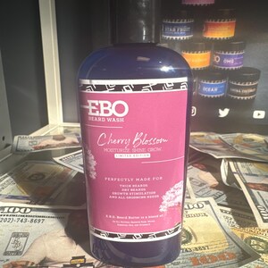 Op de afbeelding: Een blauwe fles EBO Beard Wash met de geur van kers. De fles is omringd door Amerikaanse dollarbiljetten. Het etiket zegt "Moisturize Shine Grow" en "Perfectly Made for Thick Beards, Dry Beards, Growth Stimulation and All Grooming Needs".