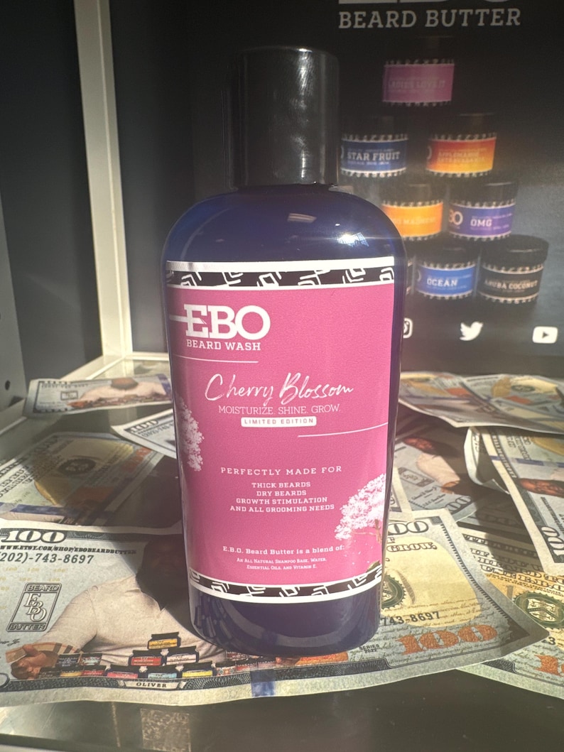 Op de afbeelding: Een blauwe fles EBO Beard Wash met de geur van kers. De fles is omringd door Amerikaanse dollarbiljetten. Het etiket zegt "Moisturize Shine Grow" en "Perfectly Made for Thick Beards, Dry Beards, Growth Stimulation and All Grooming Needs".