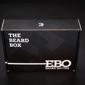 Puede incluir: Una caja negra con texto blanco que dice "THE BEARD BOX" y "EBO BEARD BUTTER". La caja tiene un diseño sencillo con un logotipo minimalista en la parte superior. El fondo es negro.