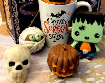 May include: A collection of Halloween-themed chocolate treats including two white ghosts, a green cauldron, a brain, a mummy, a skull, two pumpkins, and a Frankenstein figure. A white mug with the text Coffee Scream & Sugar sits among them.