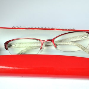 Könnte beinhalten: Rote Brillenbox mit einer roten Brille darin. Die Brille hat ein weißes, glitzerndes Design auf den Bügeln.