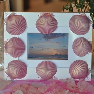 May include: A white picture frame decorated with pink seashells. The frame contains a picture of a sunset over the ocean.