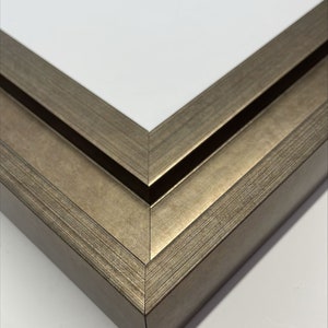 May include: Close-up of the corner of a double-layered picture frame. The frame is made of wood with a silver-toned finish. The frame has a slightly textured surface.
