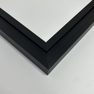 K&ouml;nnte beinhalten: Nahaufnahme einer schwarzen Bilderrahmenecke. Der Rahmen besteht aus einem dunklen, matten Material und hat ein klares, modernes Design. Die Ecke des Rahmens bildet einen scharfen 90-Grad-Winkel, mit wei&szlig;em Hintergrund.