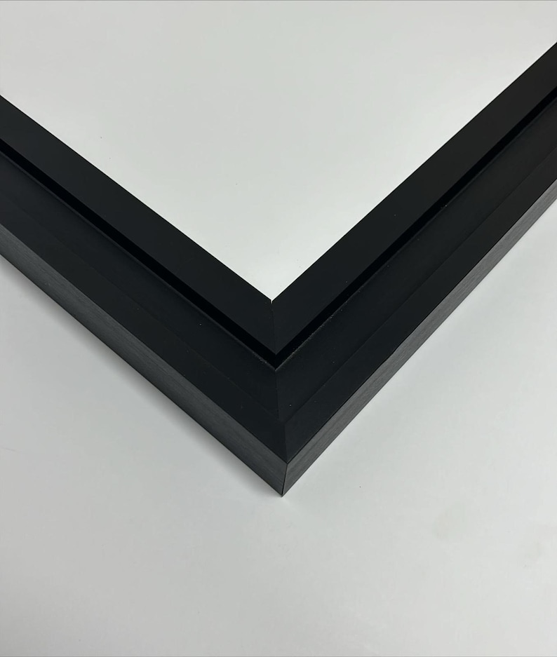 K&ouml;nnte beinhalten: Nahaufnahme einer schwarzen Bilderrahmenecke. Der Rahmen besteht aus einem dunklen, matten Material und hat ein klares, modernes Design. Die Ecke des Rahmens bildet einen scharfen 90-Grad-Winkel, mit wei&szlig;em Hintergrund.