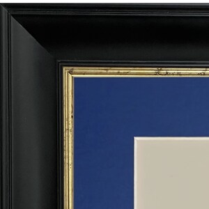 K&ouml;nnte beinhalten: Ein schwarzer Bilderrahmen mit goldener Verzierung und einem blauen Passepartout. Das Passepartout hat einen wei&szlig;en Innenrand.