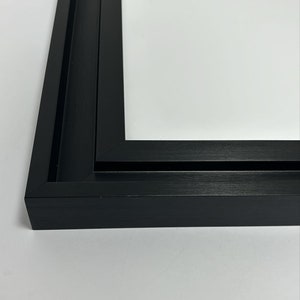 K&ouml;nnte beinhalten: Nahaufnahme einer schwarzen Bilderrahmenecke. Der Rahmen hat eine matte Oberfl&auml;che und ein schlichtes, modernes Design. Der Rahmen ist leer, mit einem wei&szlig;en Hintergrund im Inneren.