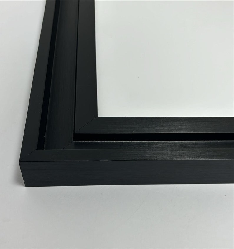 K&ouml;nnte beinhalten: Nahaufnahme einer schwarzen Bilderrahmenecke. Der Rahmen hat eine matte Oberfl&auml;che und ein schlichtes, modernes Design. Der Rahmen ist leer, mit einem wei&szlig;en Hintergrund im Inneren.