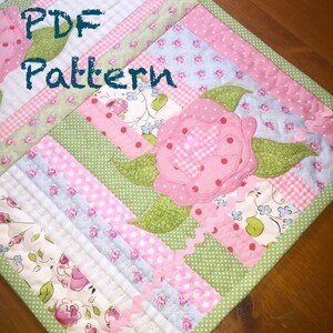 Op de afbeelding: Een PDF-patroon voor een tafelloper met een roze bloemenapplicatie en groene bladeren. De loper is gemaakt van verschillende stoffen in roze, blauw en groen met witte stippen en ruitjespatronen. De tekst "PDF Pattern" staat in het groen in de linkerbovenhoek van de loper.