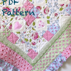 Może przedstawiać: Wzór PDF na kołdrę w patchworku z różowymi, zielonymi i niebieskimi tkaninami w kwiaty. Kołdra ma białe tło i jest ozdobiona białą koronką. Kołdra jest również ozdobiona dwoma różowymi sercami.