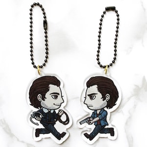 May include: Two clear acrylic keychains with black chains. Each keychain features a cartoon illustration of a man with brown hair and a blue shirt. One man is holding a gun and the other is holding a rope.