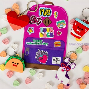 Puede incluir: Una mochila rosa con un bolsillo morado y varias pegatinas de colores. La mochila tiene el texto "I've got your back" y "Happy Valentine's Day". Hay cuatro llaveros unidos a la mochila: un corazón rojo, un taco, una taza roja con una tapa blanca y un perro morado y blanco.