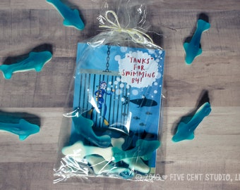 May include: A blue and white favor bag filled with blue dolphin-shaped gummies. The bag has a card attached with the text "Tanks for Swimming By!" and a cartoon illustration of a diver in a cage.