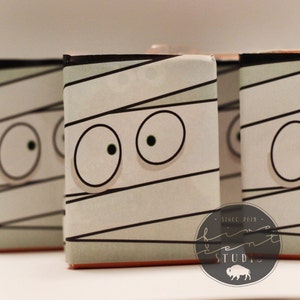 May include: Three small boxes with a mummy design. The boxes are white with black lines and have two large white eyes with green pupils. The boxes have a brown bottom and the text "Since 2015" is on the bottom right corner of the box.